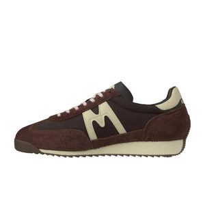 A single low-top sneaker shown in profile against a plain white background. The shoe features dark brown suede and mesh panels, a large cream-colored 'M' logo on the side, cream laces, and a thick cream midsole.