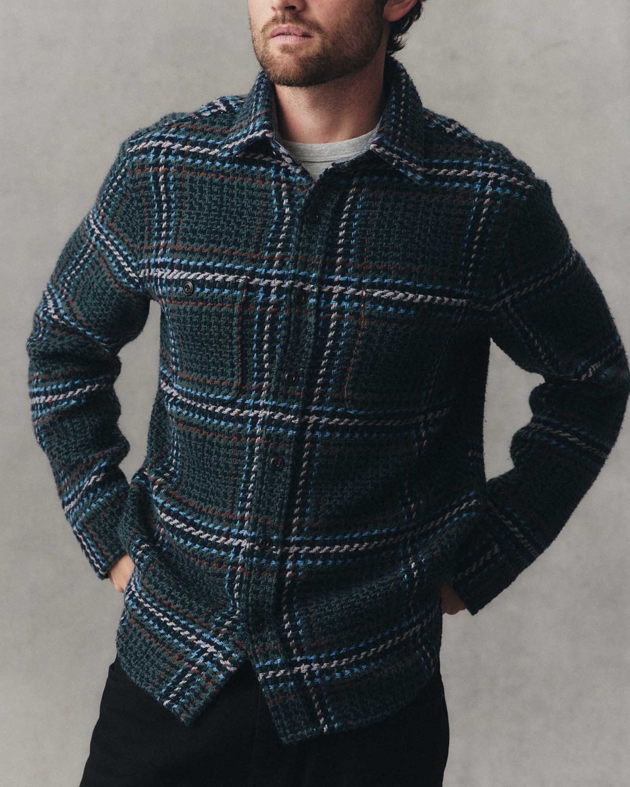 A man wearing a dark teal and black textured knit button-up shirt with a white and blue plaid pattern, paired with black pants, against a neutral gray background.
