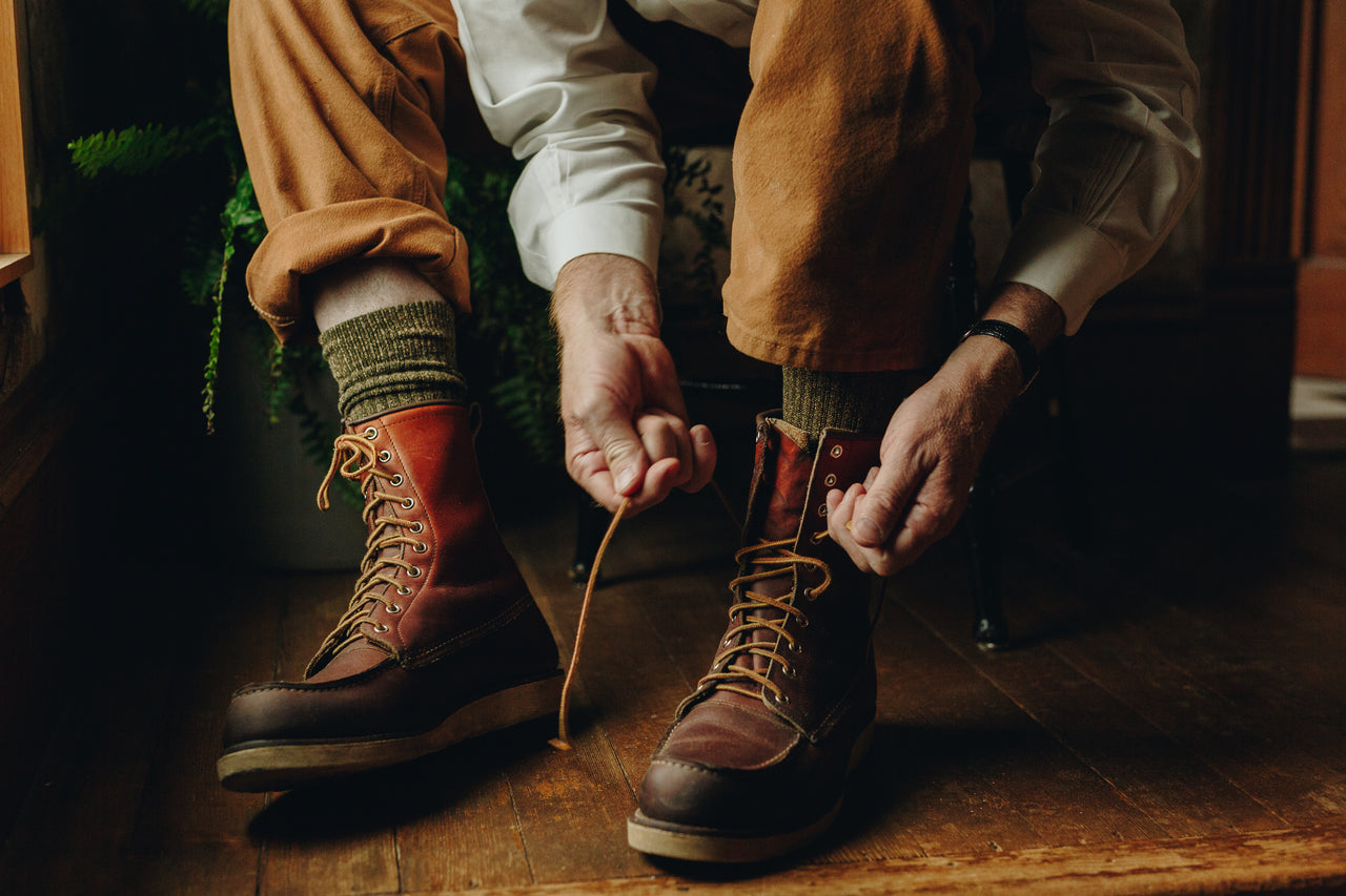 A close-up lifestyle shot of a person sitting on a wooden floor, tying the laces of a pair of rugged brown leather boots. The person is wearing brown corduroy trousers, a white long-sleeve shirt, and knit socks.