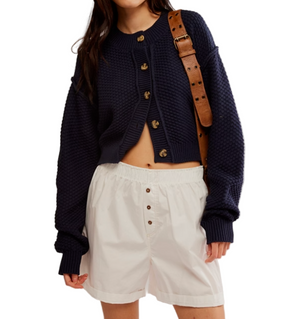 Person wearing a navy cardigan and white shorts with a brown strap over shoulder on a light gray background