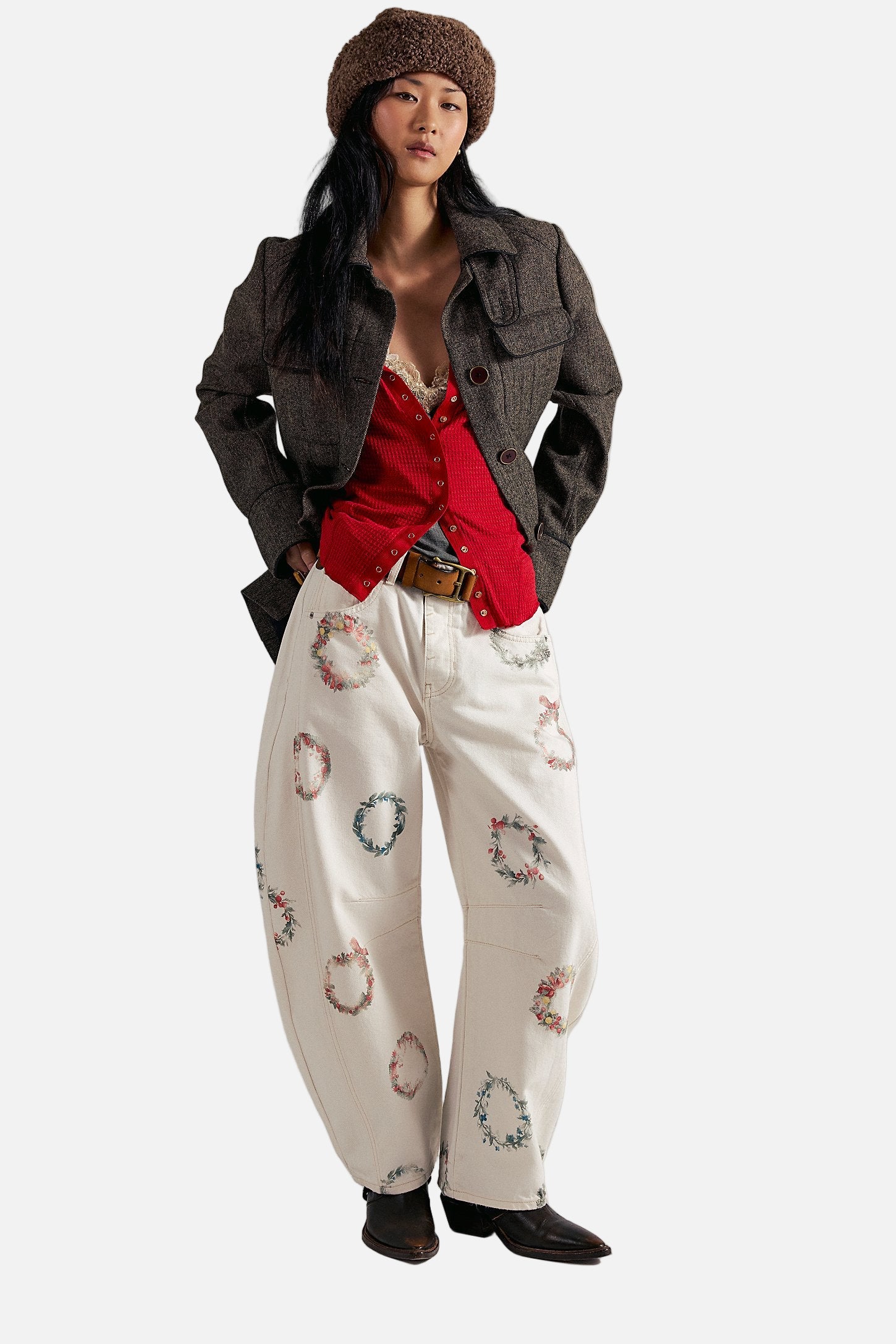 Woman wearing a red shirt, brown jacket, and white pants with floral patterns on a plain background